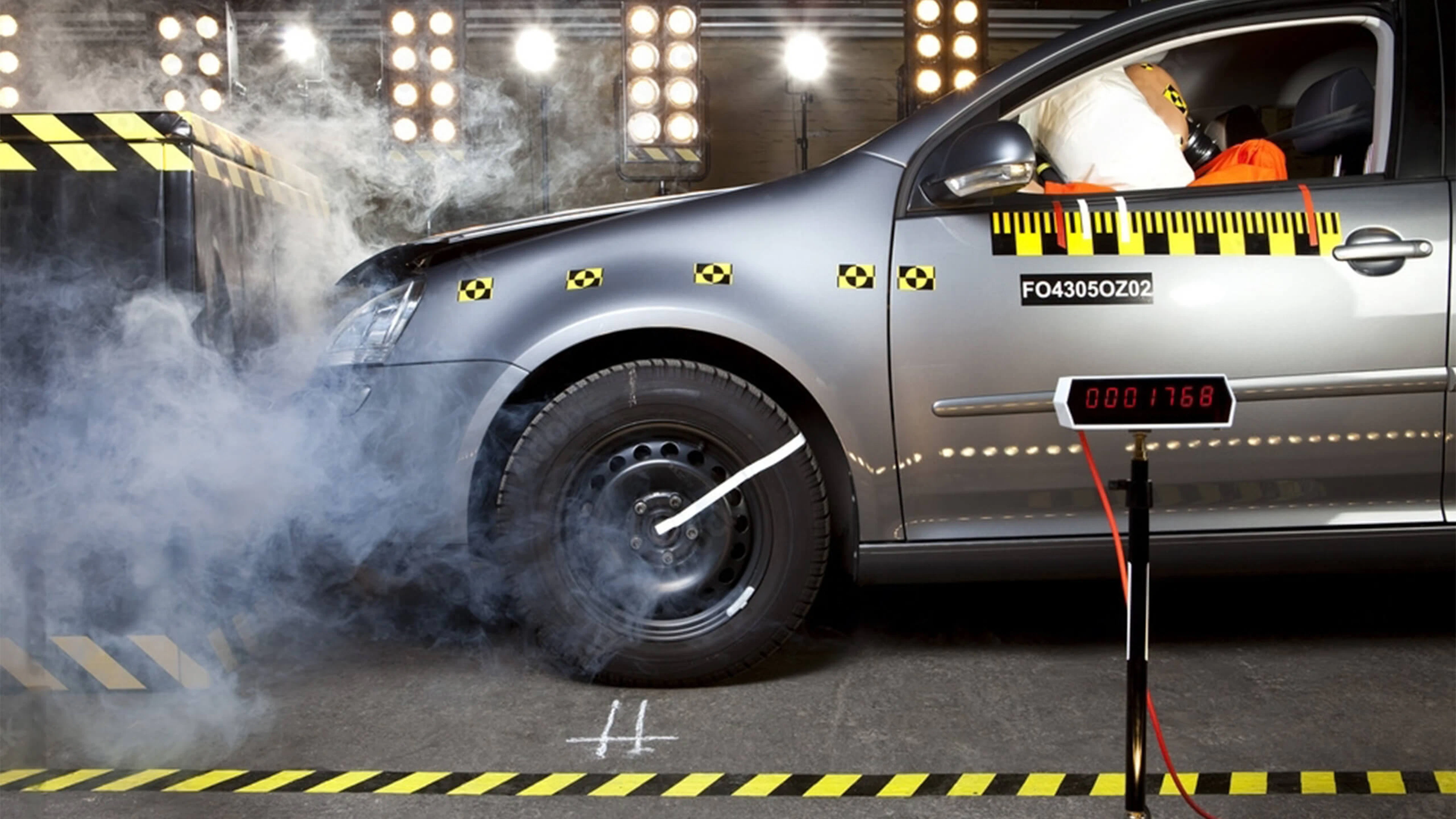 Lors d'un crash test automobile, la fumée émerge du moteur, tandis qu'un mannequin est assis au siège du conducteur, avec l'airbag déployé.
